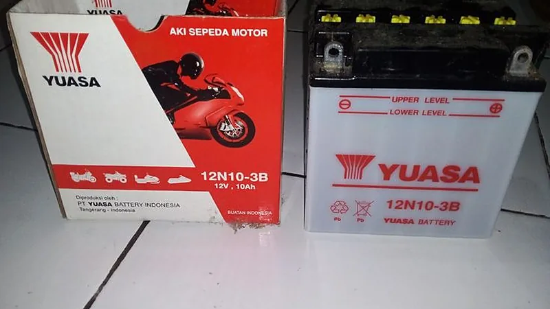 Aki basah motor Yuasa 12N10-3B buatan Indonesia bersanding dengan kardusnya, garis batas upper dan lower level terlihat di bodinya