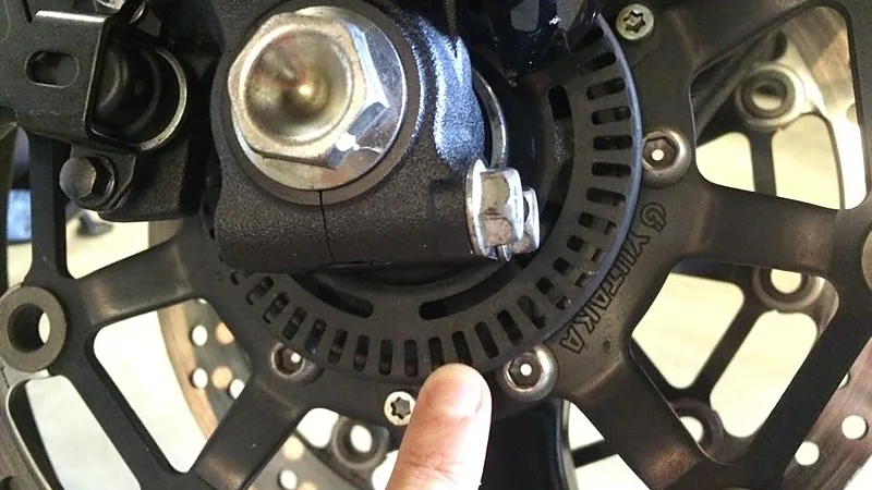 Jari menunjuk ring bergerigi sensor ABS di hub roda depan motor, dengan sensor kecepatan terpasang di atasnya