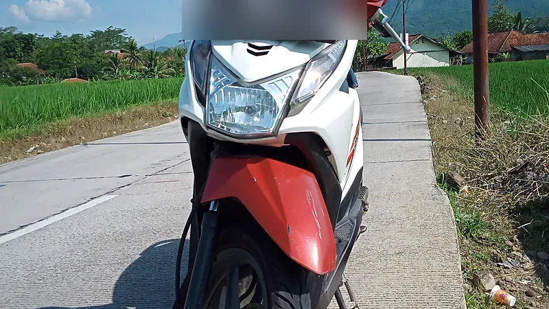 Honda BeAT putih-merah difoto dari depan di jalan desa dengan sawah di kirinya; pelat nomornya diburamkan