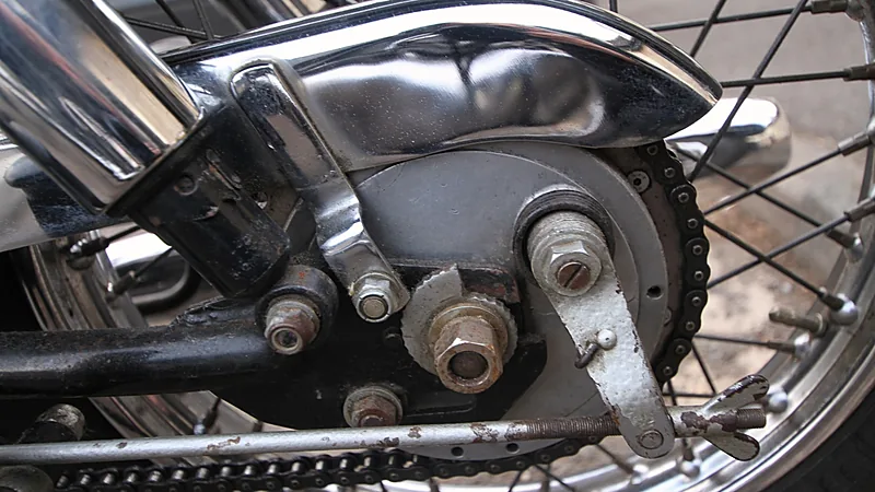 Hub roda belakang motor klasik memperlihatkan rantai, sproket, dan batang penyetel rantai pada ujung swing arm
