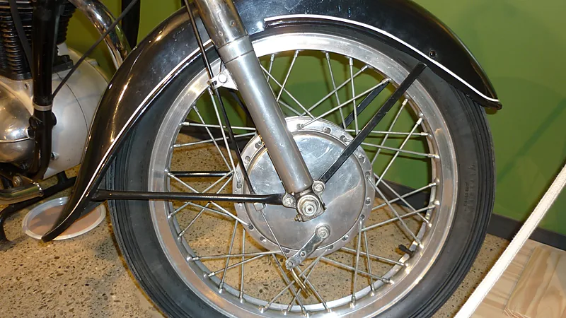 Roda jari-jari motor klasik dengan hub rem tromol besar di tengahnya, tuas dan batang penahannya terlihat jelas