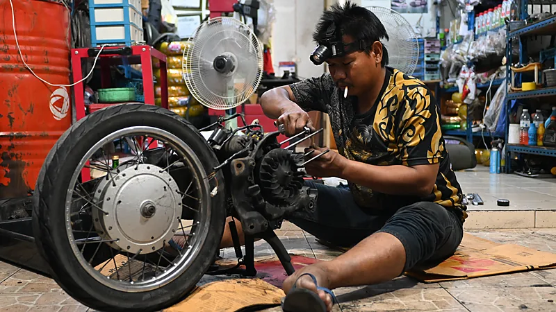 Mekanik duduk di lantai bengkel membongkar mesin motor dengan kunci di tangan, dikelilingi roda, perkakas, dan rak sparepart