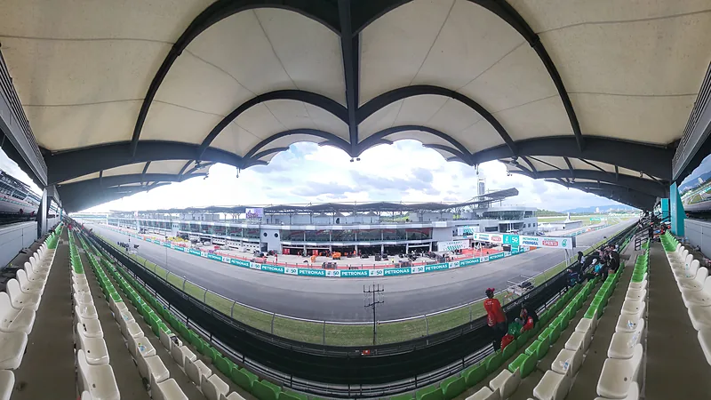 Lintasan lurus Sirkuit Sepang dilihat dari tribun beratap: pit building, papan sponsor, dan deretan kursi penonton