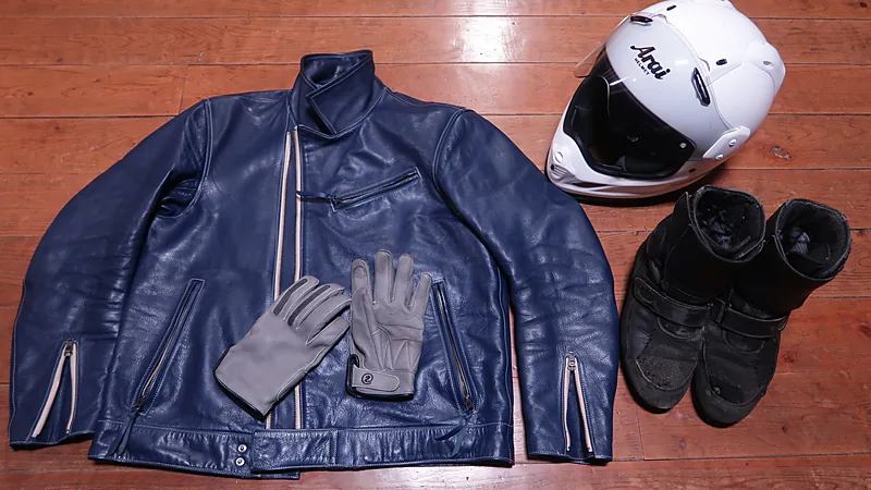 Perlengkapan berkendara lengkap tertata di lantai kayu: jaket kulit biru, helm putih Arai, sepasang sarung tangan, dan sepatu boot hitam