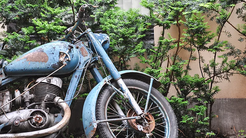 Motor klasik biru berkarat dengan cat mengelupas terparkir lama di antara semak hijau