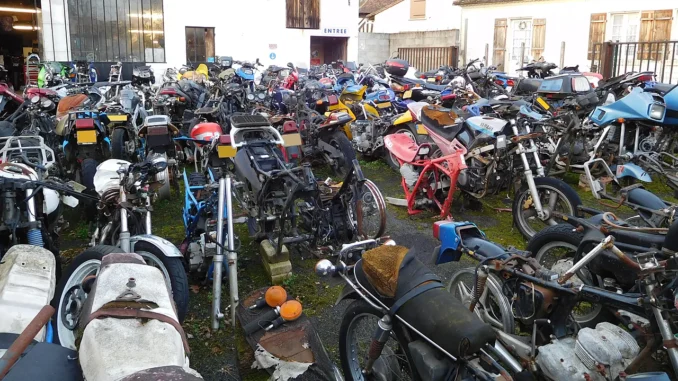 Halaman bengkel penuh motor tua yang terparkir bertumpuk dan berdebu, sebagian tinggal rangka