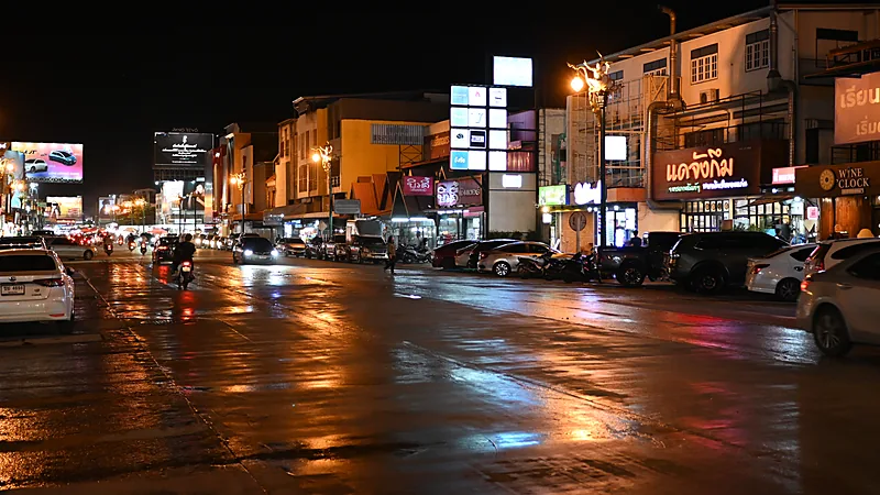 Jalan kota basah di malam hari memantulkan lampu toko dan papan nama, dengan sepeda motor melintas pelan di kiri
