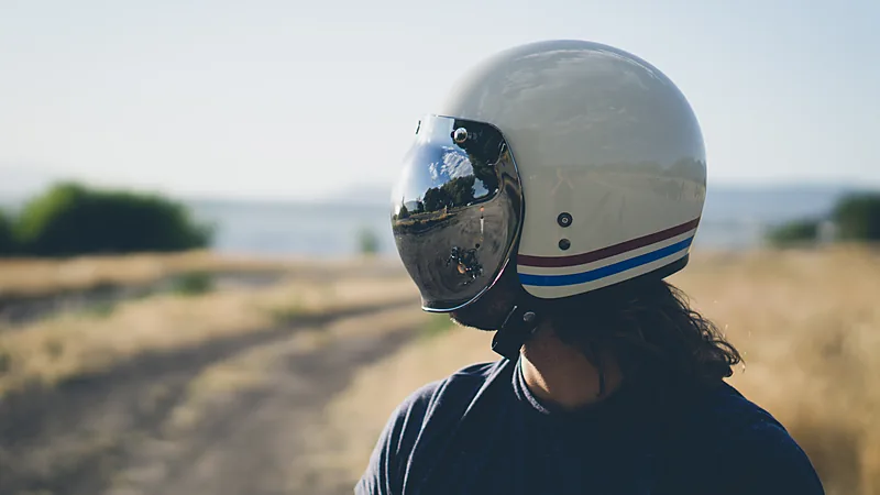 Pengendara mengenakan helm retro berwarna krem bergaris merah-biru dengan visor bubble mengilap, difoto dari belakang di jalan tanah