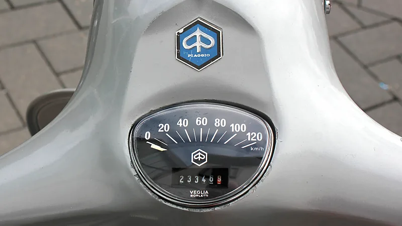 Speedometer analog Vespa bermerek Veglia Borletti dengan kaca cembung dan penunjuk odometer, terpasang di tengah setang
