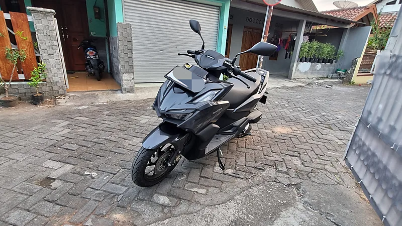 Honda Vario 160 hitam tampak depan-samping di jalan paving blok perumahan, pelat nomornya diburamkan