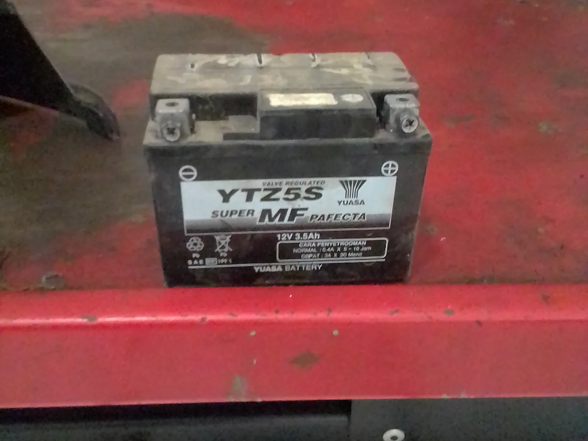 Aki kering MF motor Yuasa YTZ5S 12 volt 3,5 ampere jam tergeletak di atas meja kerja merah, label petunjuk penyetruman berbahasa Indonesia terlihat di bodinya