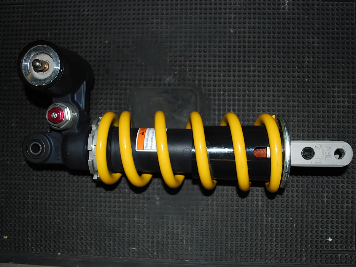 Shockbreaker monoshock terlepas di atas alas karet: per kuning, tabung reservoir di ujung atas, sekrup penyetel merah, dan ring berulir penyetel preload