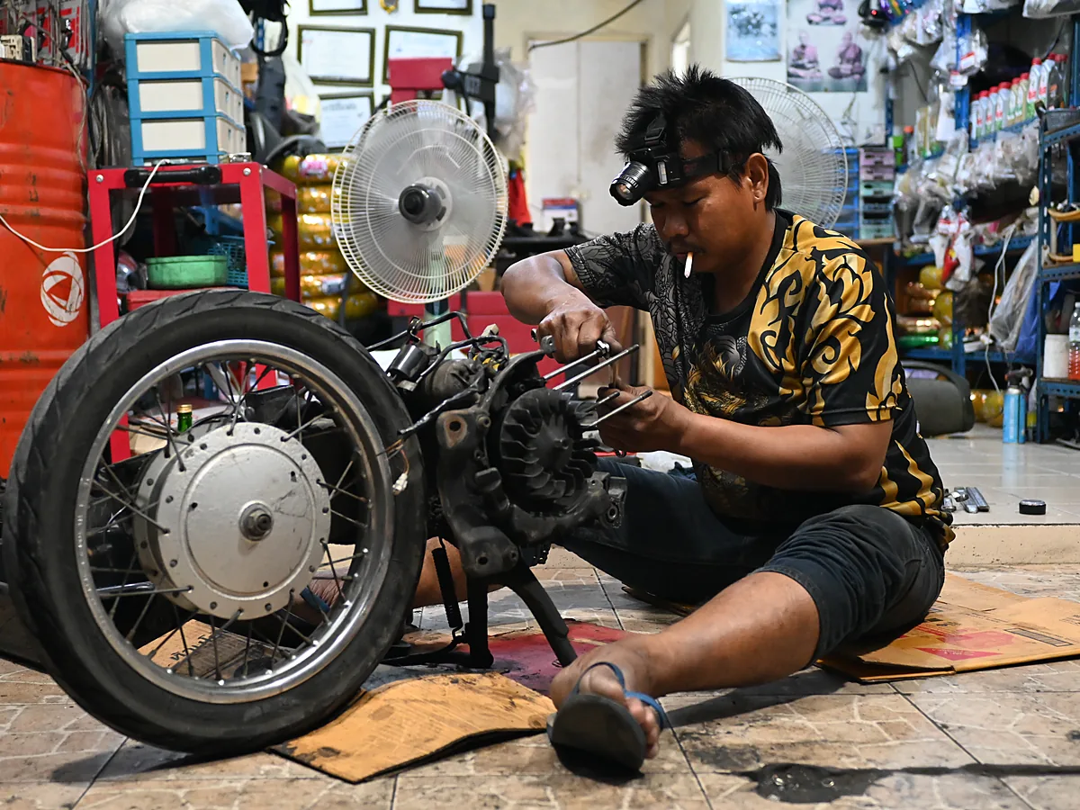 Mekanik duduk bersila di lantai bengkel membongkar mesin motor dengan kunci di tangan, dikelilingi roda, perkakas, dan rak sparepart
