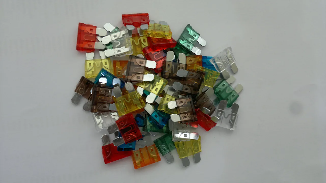 Tumpukan sekring blade berbagai warna - merah, cokelat, kuning, biru, hijau, oranye, dan bening - di atas permukaan putih