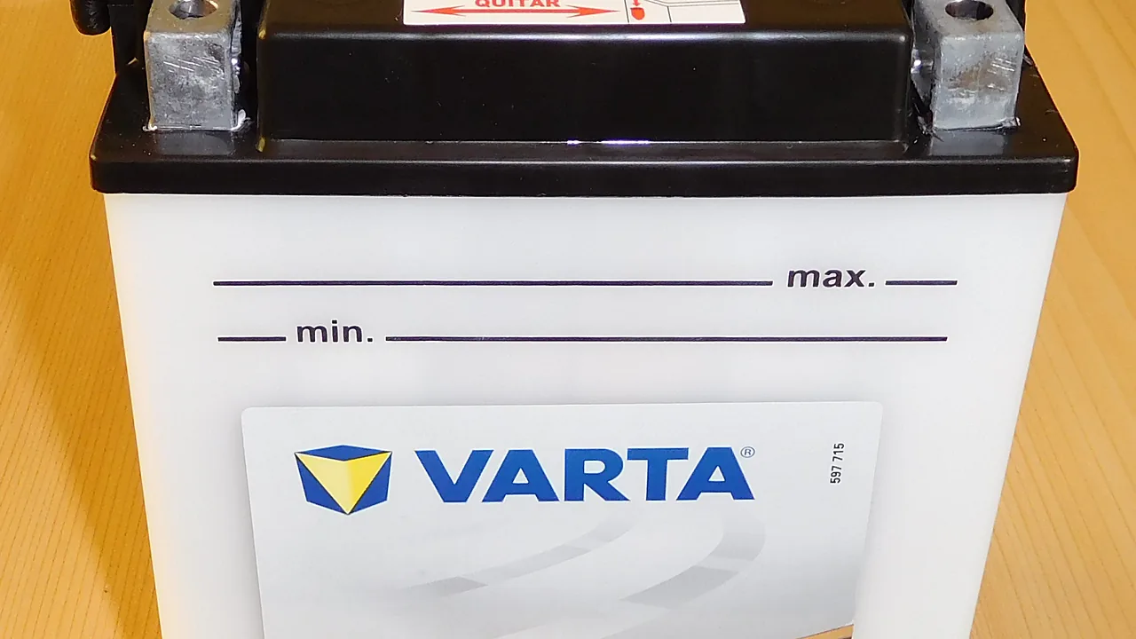 Aki motor basah merek Varta warna putih dengan dua terminal logam di atas dan garis penanda batas air min dan max di badannya